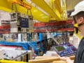 Photo of the BASE-STEP system being transported by overhead crane through the experimental hall of the Antimatter Factory at CERN. The system is an irregularly-shaped gray box and it's suspended from a large, bright yellow crane below the hall ceiling. A hard-hatted physicist, Marcel Leonhardt, looks on while holding a tablet displaying a dashboard of parameters.